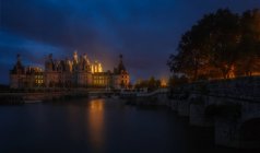 Bridge and amazing medieval castle standing on shore of river at night in Loire, France. — Stock Photo