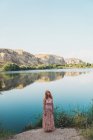 Молодая женщина в длинном платье, стоящая на скалистом берегу спокойного озера на фоне скалистых скал — стоковое фото