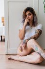 Sensual tatuado jovem mulher sentado no chão e olhando para a câmera em casa — Fotografia de Stock