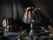 Vegan chocolate bundt cake decorated with twigs of plants on cake stand on black fabric — Stock Photo
