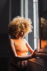 Jovem sensual mulher em roupa interior com cabelo afro em pé perto da janela com copo — Fotografia de Stock