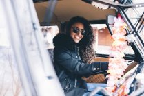 Усміхнена молода жінка в чорному пальто і сонцезахисні окуляри, сидячи всередині машини — стокове фото