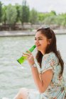 Молода усміхнена жінка п'є воду на березі річки — стокове фото