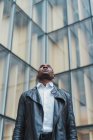 Elegante giovane uomo etnico in abiti eleganti e giacca di pelle guardando contro l'edificio in vetro moderno — Foto stock