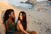 Lächelnde multiethnische Frauen, die sich an der Küste im Sonnenlicht umarmen und chillen — Stockfoto