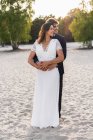 Amare l'uomo abbracciare bella sposa in abito elegante e guardarsi mentre in piedi sulla costa sabbiosa alla luce del sole — Foto stock