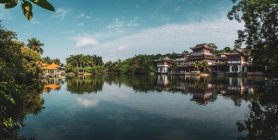 Aguas tranquilas lago que refleja el cielo y los edificios orientales en la orilla con exuberante vegetación tropical, Qingxiu Montaña, China - foto de stock