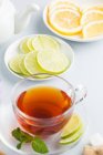 Black tea in glass cup with brown sugar, cinnamon, mint and slices of citrus fruits on saucers with teapot on white background — Stock Photo