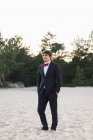 Erwachsener Mann trägt eleganten schwarzen Anzug mit Fliege und steht am Strand mit den Händen in den Taschen und blickt in die Kamera — Stockfoto