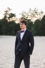 Homem adulto vestindo elegante terno preto com gravata borboleta e em pé na praia com as mãos nos bolsos olhando para a câmera — Fotografia de Stock