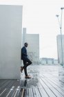 Афроамериканский бизнесмен опирается на стену на открытом воздухе с современными зданиями на заднем плане — стоковое фото