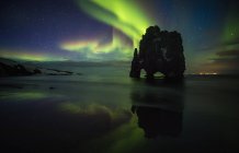 Скалистая скала, стоящая в море на фоне яркого северного сияния в Исландии. — стоковое фото