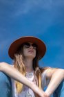 De baixo tiro de jovem mulher confiante com cabelos longos vestindo trajes casuais com óculos de sol e chapéu sentado sob o céu azul — Fotografia de Stock