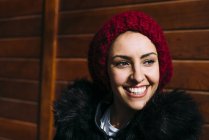Sonriente mujer joven en sombrero de lana roja de pie sobre fondo de madera y mirando hacia otro lado - foto de stock