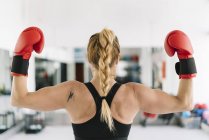 Вид на мускулистую женщину в красных боксёрских перчатках, показывающих бицепсы, стоя на размытом фоне спортзала — стоковое фото