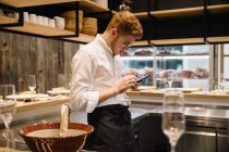 Vista laterale del giovane in uniforme cuoco appoggiato sul bancone della cucina e utilizzando smartphone moderno mentre in piedi in cucina ristorante — Foto stock