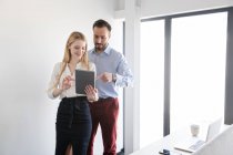 Homem e mulher sorridentes elegantes em pé no escritório leve e usando tablet juntos durante a discussão — Fotografia de Stock