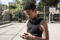 Menino afro ouvindo música com smartphone e fones de ouvido na quadra de basquete — Fotografia de Stock