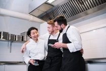 Drei unterschiedliche Männer in Kochuniform lachen und surfen auf Smartphones, während sie gemeinsam in der Restaurantküche stehen — Stockfoto