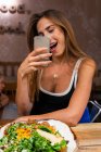 Счастливая молодая женщина сидит за деревянным столом с миской еды и фотографируется со смартфоном — стоковое фото