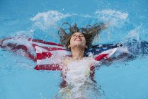 Relaxado sorrindo mulher espirrando na piscina com bandeira americana — Fotografia de Stock