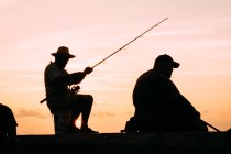 Black silhouette of men fishing on seafront against light sunset sky, Cuba. — Stock Photo