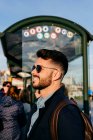 Вид збоку модного бородатого чоловіка в сонцезахисних окулярах, що стоїть з рюкзаком на сонячному світлі на залізничній станції і посміхається . — стокове фото