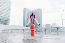 Strong ethnic man in sportswear jumping over red obstacle on wet pavement with modern city — Stock Photo