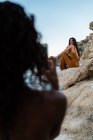 Чорна жінка фотографує зі смартфоном стильного друга, який сидить на скелястій скелі приморського узбережжя влітку — стокове фото