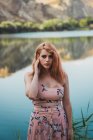 Mulher sonhadora em vestido de verão em pé na margem do lago e olhando para a câmera — Fotografia de Stock