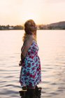 Молодая блондинка с мокрыми волосами в летнем платье, стоящая в воде озера на закате — стоковое фото