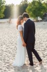Любящий мужчина обнимает красивую невесту в элегантном платье и смотрит друг на друга, стоя на песчаном побережье под солнечным светом — стоковое фото