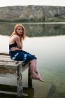 Sinnliche junge Frau im Kleid sitzt auf einem Steg am See in der Natur — Stockfoto