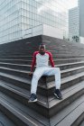 Jeune homme ethnique en vêtements de sport assis sur les escaliers et contre les bâtiments en verre dans la ville — Photo de stock