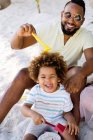 Homem étnico adulto alegre em óculos de sol sentado na areia e brincando com o filho em férias — Fotografia de Stock
