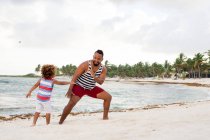 Cheerful African-American man playing and having fun with son at the ocean on sandy beach — Stock Photo
