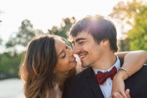 Heureux homme et femme élégants dans des tenues de mariage embrassant sur le rocher de la plage et regardant souriant loin dans la lumière du soleil — Photo de stock