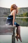 Träumende zarte Frau im Kleid mit geschlossenen Augen sitzt auf einem Holzsteg über dem Wasser des Sees — Stockfoto