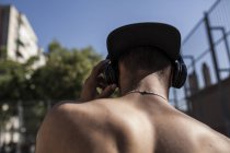 Вид сзади на мальчика без рубашки в кепке, слушающего музыку с наушниками на улице — стоковое фото