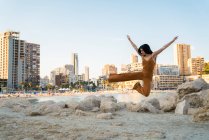 Игривая женщина в стиле в целом делает расколы на берегу со зданиями на заднем плане — стоковое фото