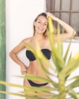 Приваблива молода леді в чорному купальнику посміхається і подряпає голову, стоячи біля будинку на курорті — стокове фото