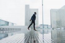 Homem étnico em roupas de negócios elegantes pulando no pavimento molhado com vidro da cidade e edifícios de concreto no fundo — Fotografia de Stock