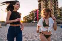 Laughing female friends running on beach with buildings on background — Stock Photo