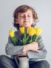Мальчик с закрытыми глазами сидит и обнимает кучу желтых тюльпанов на сером фоне — стоковое фото