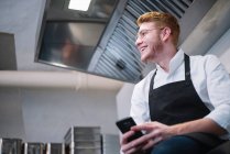Вид збоку молодого чоловіка в кулінарному уніформі, що спирається на кухонну стійку і використовує сучасний смартфон, стоячи на кухні ресторану — стокове фото