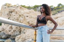 Dreamy young ethnic woman in lace top and high-waist jeans standing and leaning on fence against rocky coastal cliffs — Stock Photo
