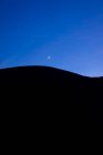 Минималистский пейзаж черного силуэта горных холмов против сумеречного голубого неба с полумесяцем — стоковое фото