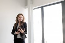 Елегантна жінка в костюмі і використовує планшет, стоячи в яскравому денному світлі всередині сучасного офісу — стокове фото