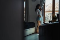 Мечтательная женщина в рубашке и шортах, стоящая у окна — стоковое фото