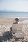 Молодая женщина сидит с напитком и смотрит на шезлонг на пляже — стоковое фото
